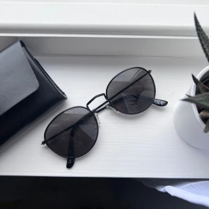 Svarta runda solglasögon - Snygga svarta solglasögon med rundade glas och tunn metallbåge. De har mörka linser och näskuddar. Kommer med ett stilrent svart fodral som håller dem skyddade när du inte använder dem samt en putsduk. Önskar du veta exakta mått? Tveka inte att höra av dig!