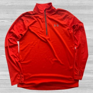 Under Armour 1/3 Zip - Sportig röd långärmad träningströja från Under Armour med svart half zip och meshpaneler för extra ventilation. Tröjan har diskret mönster på axlarna och är tillverkad i lätt polyester som andas. Perfekt för löpning eller gymmet.