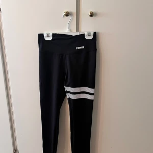 Svarta leggings från Stronger XS - Svarta träningstights från Stronger i storlek XS med två vita ränder på ena benet. Hög midja och tight passform, perfekta för träning. Använd vid fåtal tillfällen och har inga defekter. 