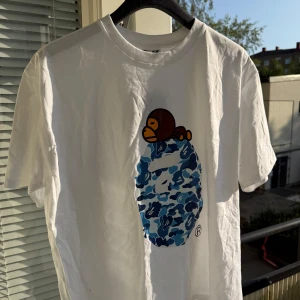 Vit BAPE t-shirt med blå camo print - Säljer en clean vit t-shirt från BAPE med ikoniskt blått camo-tryck och en apa på bröstet. Klassisk passform och tillverkad i mjuk bomull. Perfekt för dig som gillar streetwear och vill sticka ut med en unik design.