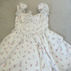 Vit blommig kortklänning med puffärm - Supersöt vit kortklänning med rosa blommor och gröna blad, puffärmar och volangdetaljer. Klänningen har en romantisk vibe med omlottkänsla upptill och små rosetter. Perfekt för sommarens alla tillfällen!