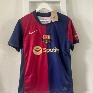 FC Barcelona Lamine Yamal tröja Nike S - FC Barcelona matchtröja från Nike i storlek S, med Lamine Yamal och nummer 19 på ryggen. Tröjan är delad i rött och mörkblått, har korta ärmar och guldiga detaljer som klubbmärke, sponsorer och tryck. Tillverkad i lätt och ventilerande polyester.