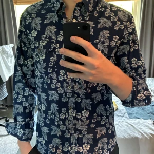 Blommig blå skjorta från Hugo Boss - Hej, säljer en snygg blå skjorta från Hugo Boss med vitt blommigt mönster och två bröstfickor. Slim fit-modell med långa ärmar och klassisk krage. Perfekt för dig som vill sticka ut med en cool och somrig vibe. Skjortan är i mjuk bomull och har knäppning framtill. Hör av dig vid frågor eller funderingar!😁