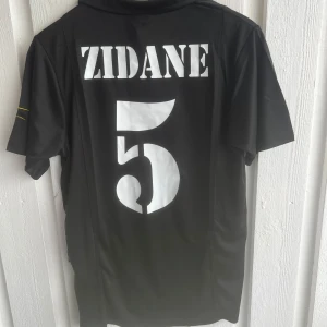 Real Madrid Zidane svart fotbollströja - Svart Real Madrid fotbollströja från Adidas med tryck 'ZIDANE' och nummer 5 på ryggen. Tröjan har klubbmärke och Adidas-logga på bröstet, samt krage och korta ärmar. Materialet är lätt och andas, perfekt för fotboll eller träning.