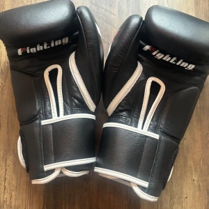 Svarta boxningshandskar från Fighting - Säljer ett par svarta boxningshandskar från Fighting med vita detaljer och logga. Handskarna är tillverkade i skinn och har breda kardborreband för extra stöd runt handleden. Perfekta för träning och sparring. Snygg och klassisk design.