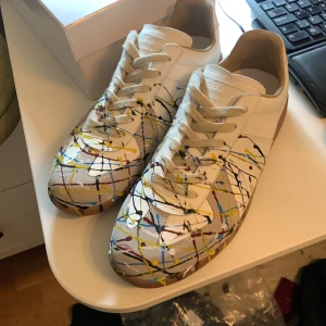 Maison Margiela sneakers med färgstänk - Unika vita sneakers från Maison Margiela med coola färgstänk i blått, gult, rött och svart. Skorna har snörning, beige detaljer och gummisula. Tillverkade i skinn och har en klassisk rund tå. Perfekta för dig som vill sticka ut med din stil.
