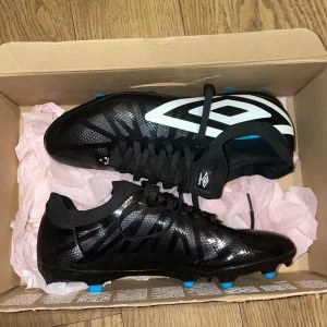 Svarta fotbollsskor från Umbro - Snygga svarta fotbollsskor från Umbro med vita detaljer och blå dobbar. Skorna har snörning och en modern design som passar perfekt för fotbollsplanen.