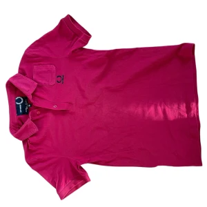 Röd pikétröja från Raf Simons - Snygg röd pikétröja från Raf Simons med klassisk krage och knappslå. Perfekt för en stilren look. Tröjan har en liten bröstficka och broderad logga på bröstet. Passar perfekt för både vardag och mer avslappnade tillfällen.