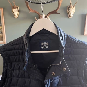 Svart quiltad väst från Boggi - Snygg svart quiltad väst från Boggi med dragkedja och knappar. Perfekt för lager-på-lager under kyliga dagar, funkar även perfekt nu på vårkvällar eller till skolan. Klassisk design med en stilren look.