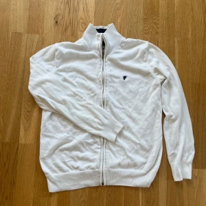 Vit kofta från Denim Culture - Säljer en stilren vit kofta från Denim Culture i storlek M. Koftan är tillverkad i 100% bomull och har en dragkedja framtill. Den har en broderad logga på bröstet och ribbade muddar. Perfekt för en avslappnad look.