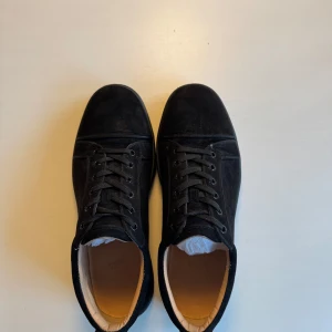 Christian Louboutin skor - Snygga svarta sneakers från Christian Louboutin med den ikoniska röda sulan i storlek 43. Skorna har snörning och är tillverkade i mocka, vilket ger en lyxig känsla. Perfekta för att ge din outfit en exklusiv touch. Allt og tillkommer, skicket är inte perfekt och därför priset. Hör gärna av er vid frågor😉