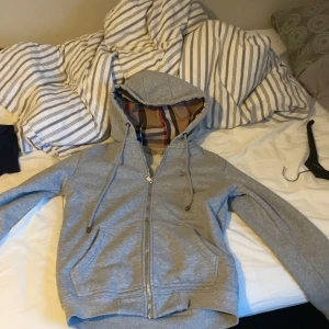 Grå hoodie med dragkedja - Säljer en grå hoodie med dragkedja och randigt foder i huvan. Den har långa ärmar och snören vid huvan för justering. Perfekt för en avslappnad stil.