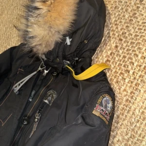 Svart parkajacka från Parajumpers - Säljer en svart parkajacka från Parajumpers med pälsfodrad huva och gul detalj vid halsen. Jackan har flera fickor och en dragkedja framtill. Perfekt för kalla dagar!