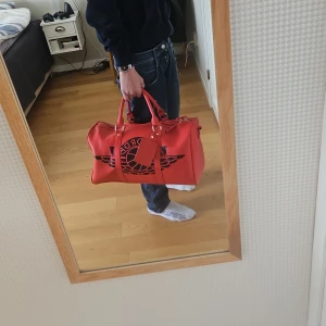 Röd väska från Jordan - Säljer en snygg röd väska från Jordan med ett stort svart logotyptryck. Väskan har både handtag och en justerbar axelrem för enkel bärning. Den är utrustad med en dragkedja upptill och har guldfärgade detaljer. Perfekt för att bära med sig det nödvändigaste med stil.