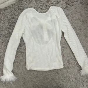 Vit ribbad topp med fjäderdetaljer - Säljer en stilren vit ribbad topp med långa ärmar och fjäderdetaljer vid ärmsluten. Toppen har en öppen rygg med knytning upptill, vilket ger en elegant touch. Perfekt för att lyfta din outfit till nästa nivå! Helt oanvänd!🦋
