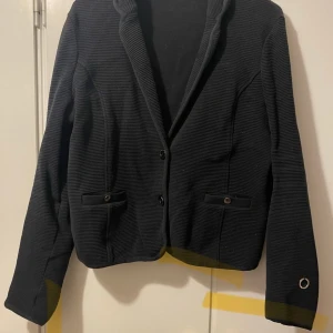 Svart kavaj från Stockholm  - Snygg svart kavaj från Stockholm  med knappar framtill och ribbad textur. Kavajen har långa ärmar och två fickor med knappdetaljer. Perfekt för en stilren look.