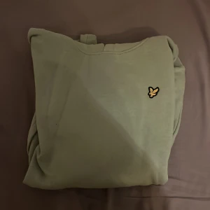 Grön hoodie från Lyle & Scott - Säljer en stilren grön hoodie från Lyle & Scott med deras ikoniska logga på bröstet. Tröjan har en klassisk känguruficka och ribbade muddar vid ärmslut och nederkant. Perfekt för en avslappnad stil.