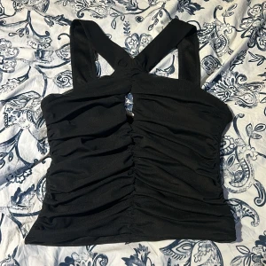 Svart topp från Gina Tricot - Snygg svart topp från Gina Tricot med breda axelband och rynkade detaljer framtill. Perfekt för en stilren look. Toppen har en smickrande passform och är tillverkad i ett mjukt material och väldigt stretchig