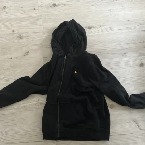 Svart hoodie från Lyle & Scott - Säljer en svart hoodie från Lyle & Scott Junior. Den har en dragkedja framtill och en liten gul logga på bröstet. Perfekt för en avslappnad stil med långa ärmar och en bekväm huva. Pris kan diskuteras via dm