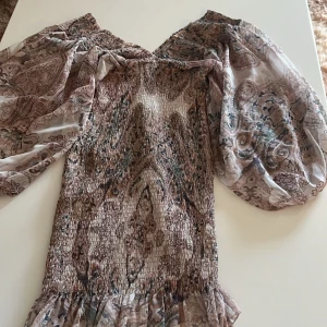 Mönstrad offshoulder-top - Säljer en snygg offshoulder-klänning som är tajt , utsväng i slutet med ett unikt mönster i jordnära toner. Klänning har långa, puffiga ärmar och en smockad kropp som ger en fin passform. Perfekt för att ge en bohemisk touch till din outfit.