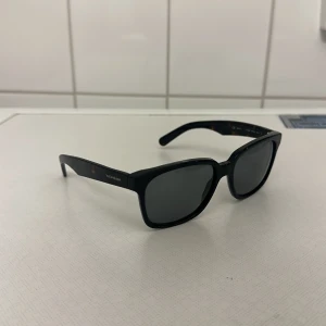 Yves Saint Laurent solglasögon  - Snygga svarta solglasögon från Yves Saint Laurent med en klassisk rektangulär form. Perfekta för att ge en stilren look. Märkesloggan syns tydligt på skalmen.