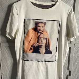 Vit t-shirt med tryck från Limitato - Säljer en vit t-shirt från Limitato med ett unikt tryck av två personer i färg. T-shirten har en normal passform och är kortärmad, perfekt för en avslappnad stil. Tillverkad i Portugal. nackdelen är att det finns en gul fläck i varje armhåla av tröjan därför den säljs av det priset den gör.