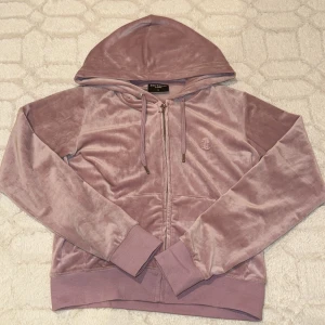 Ljusrosa hoodie från Juicy Couture - Säljer en mjuk och stilren ljusrosa hoodie från Juicy Couture. Den har en dragkedja framtill och en broderad logga på bröstet. Perfekt för en avslappnad look med sin sköna passform och huva.