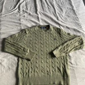 Grön stickad tröja från Ralph Lauren - Säljer en stilren grön stickad tröja från Ralph Lauren med kabelstickat mönster och broderad logga på bröstet. Tröjan har långa ärmar och ribbstickade muddar vid ärmslut och nederkant. Perfekt för kyliga dagar!