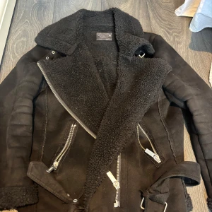 Svart jacka från Crocker - Snygg svart jacka från Crocker med dragkedjor och bälte. Jackan har en mysig foder och är perfekt för kyligare dagar. Den har långa ärmar och en cool look med flera detaljer.