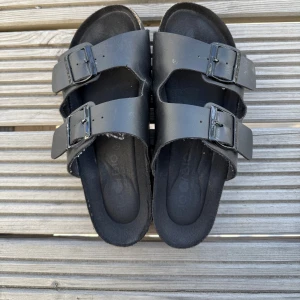 Svarta sandaler med spännen - Säljer ett par svarta sandaler med dubbla justerbara spännen. Perfekta för sommardagar och ger en avslappnad stil. Sandalerna har en bekväm fotbädd och är lätta att bära.