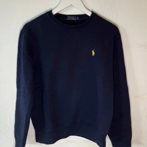 Ralph Lauren sweatshirt - Mörkblå sweatshirt från Ralph Lauren med klassisk gul broderad logga. Använd endast några gånger och är i väldigt fint skick. Tröjan är stilren, prefekt för vardagen men även lite mer uppklätt. Hör av dig vid intresse!