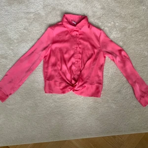 Rosa blus med knyt - Snygg rosa blus med knyt framtill och långa ärmar. Perfekt för en chic look. Blusen har knappar hela vägen och en klassisk krage.