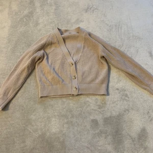 Beige ribbad kofta med knappar - Säljer en stilren beige kofta med ribbad struktur och knappar framtill. Koftan har långa ärmar och en v-ringad design, perfekt för lager-på-lager-stil. Passar till både vardag och mer uppklädda tillfällen.