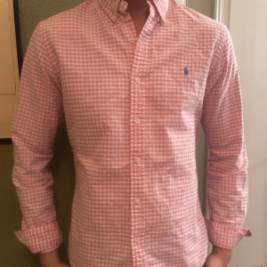 Rosa rutig skjorta från Ralph Lauren - Snygg rosa rutig skjorta från Ralph Lauren med klassisk krage och knappar framtill. Den har en broderad logga på bröstet och långa ärmar. Perfekt för en stilren look. Perfekt till sommaren!