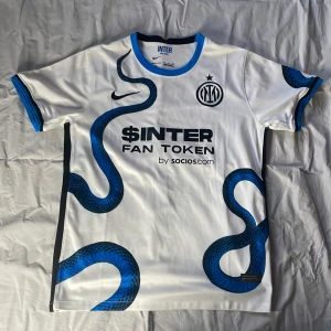 Vit och blå Inter fotbollströja - Säljer en vit och blå Inter fotbollströja från Nike med ett unikt ormmönster. Tröjan har korta ärmar och klubbens logga på bröstet. Perfekt för fans som vill visa sitt stöd med stil! 🐍