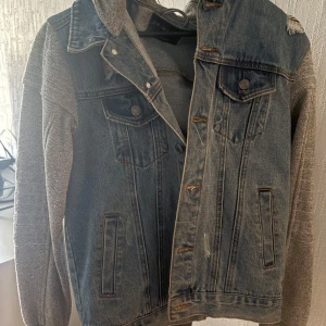 Jeansjacka med grå ärmar - Snygg jeansjacka från Bershka med grå stickade ärmar och krage. Jackan har knappar framtill och två bröstfickor med lock. Perfekt för en casual look!