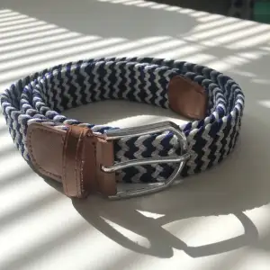 Snyggt randigt bälte i blått och vitt med bruna läderdetaljer. Bältet har en klassisk metallspänne och är perfekt för att ge en stilren touch till din outfit.