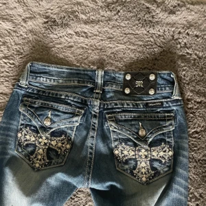 Miss Me blå straight jeans med broderi - Snygga blå jeans från Miss Me med straight fit och coola broderade detaljer på bakfickorna. Fickorna har korsmönster och glittriga knappar. Jeansen har slitningar och tvättad look, lowwaist och klassisk femficksdesign. Perfekt för dig som gillar statement-plagg. Midjemåttet är 35 cm och innerbenet är 72 cm🥰