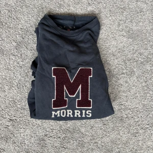 Mörkblå sweatshirt från Morris - Snygg mörkblå sweatshirt från Morris med stor vinröd M i frotté på bröstet och broderad text. Klassisk collegekänsla med ribbade muddar och rund hals. Perfekt för dig som gillar stilren och sportig look.