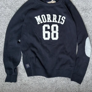Svart stickad tröja Morris Gents - Svart stickad tröja från Morris Gents med vit text 'MORRIS 68' på bröstet och ljusgråa armbågslappar. Tröjan har ribbade muddar och rund hals. Perfekt för dig som gillar sportig och klassisk stil.