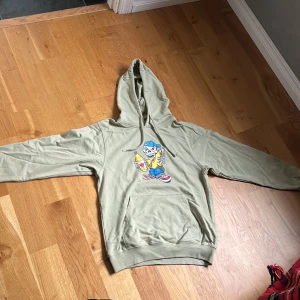 Olivgrön hoodie med från Vans - Säljer en beige hoodie från Vans i loose fit, storlek XS. Tröjan har ett stort färgglatt tryck med en tecknad figur på framsidan, huva med snörning och en klassisk magficka. Perfekt för dig som gillar streetwear och chill stil. Jag har aldrig använt den här tröjan den har endast legat i min garderob.