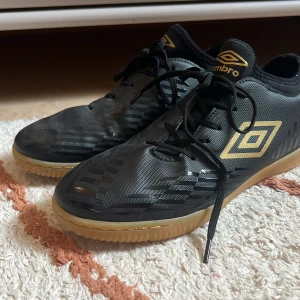 Svarta och guldiga Umbro fotbollsskor - Säljer ett par svarta Umbro Raposa fotbollsskor med guldiga detaljer och logga. Skorna har snörning, mönstrad ovandel och gummisula i beige. Perfekta för inomhusfotboll och har en sportig look som sticker ut på planen. Kan tänka mig att sänka priset vid snabb affär!