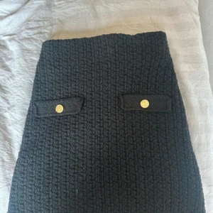 Svart stickad kortkjol från H&M - Svart stickad kortkjol från H&M med grov struktur och två dekorativa ficklock framtill med guldiga knappar. Kjolen har en klassisk rak passform och är perfekt för dig som gillar en stilren och trendig look.