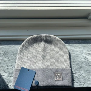 Grå rutig mössa Louis Vuitton - Snygg grå mössa från Louis Vuitton med diskret rutigt mönster och uppvikt kant. LV-logga framtill och exklusiv känsla. Perfekt accessoar för dig som gillar stilrena designer och vill sticka ut med ett lyxigt märke. Materialet är mjukt och håller dig varm.
