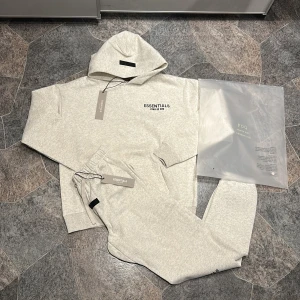 Essentials Fear of God hoodie ljusgrå S - Ljusgrå hoodie från Essentials Fear of God i storlek S. Klassisk oversized passform med huva och svart logga tryckt på bröstet och ryggen. Tillverkad i mjuk bomullsblandning, perfekt för chill dagar. Snygg och enkel stil som funkar året runt.