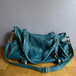 Blå axelväska - genuine leather - Snygg blå axelväska i äkta skinn med justerbar axelrem och guldfärgade metalldetaljer. Väskan har dragkedja upptill, flera innerfack och coola knytband på sidorna. Perfekt storlek för allt du behöver ha med dig. Tecken på användning finns men den är fortfarande väldigt fin.