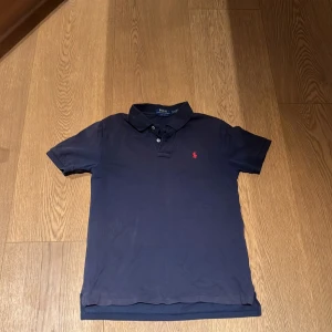 Mörkblå piké från Polo Ralph Lauren - Klassisk mörkblå pikétröja från Polo Ralph Lauren med korta ärmar och krage i strorleken junior L 14-16. Tröjan har två knappar vid halsen och den ikoniska röda loggan broderad på bröstet.