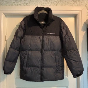 Svart pufferjacka från Sail Racing - Säljer en svart pufferjacka från Sail Racing med hög krage och dragkedja framtill. Jackan är quiltad och har en stilren look med logga på bröstet och ryggen. Perfekt för kalla dagar när du vill hålla värmen och ändå se snygg ut. En reva på ryggen finns, därav priset. Se sista bild. 