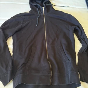 Svart hoodie Peak Performance L - Svart hoodie från Peak Performance i storlek L. Klassisk modell med dragkedja, huva med snörning och två stora fickor framtill. Perfekt för chill dagar eller när du vill ha en enkel och stilren look.