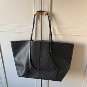 Svart shoppingväska från By Malene Birger - Stilren och rymlig shoppingväska från By Malene Birger i svart/brun med diskret rutigt mönster. Väskan har långa svarta axelband och beige insida med en stor innerficka. Perfekt för dig som vill ha en snygg och praktisk väska till vardagen. Den är knappt använd ❤️